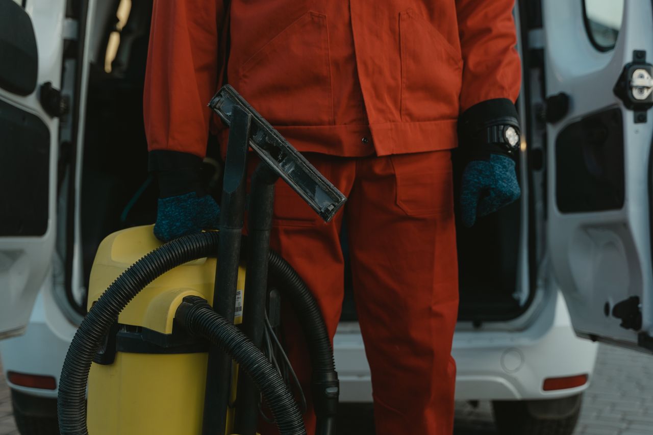 Un nettoyeur professionnel en uniforme rouge tenant un aspirateur près d'un van, prêt à intervenir.